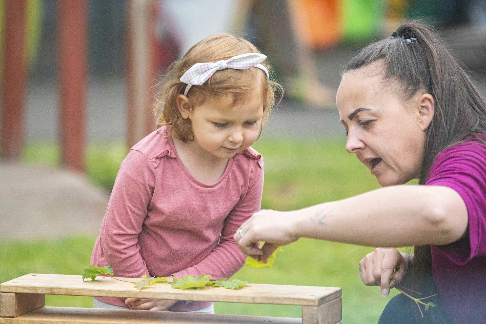 Female nursery worker showing a little girl leaves and creatures in their outdoor garden at Pelsall Village Day Nursery