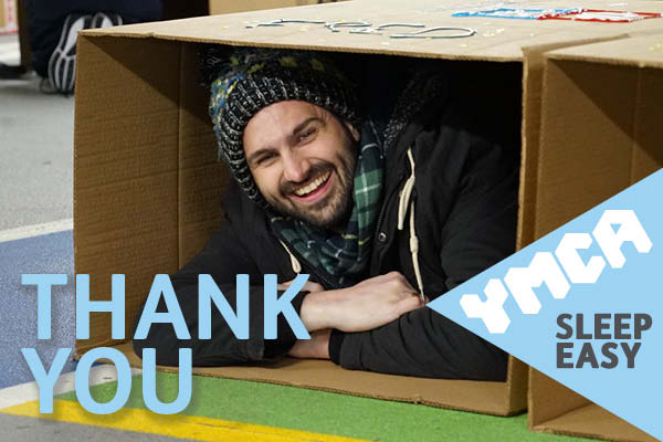 A young man wearing a wooly hat is lying inside a cardboard box smiling.