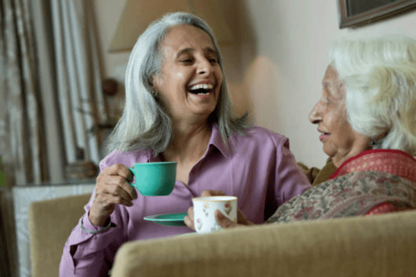 An older lady with her friend enjoying a cup of tea together, laughing of the sofa.