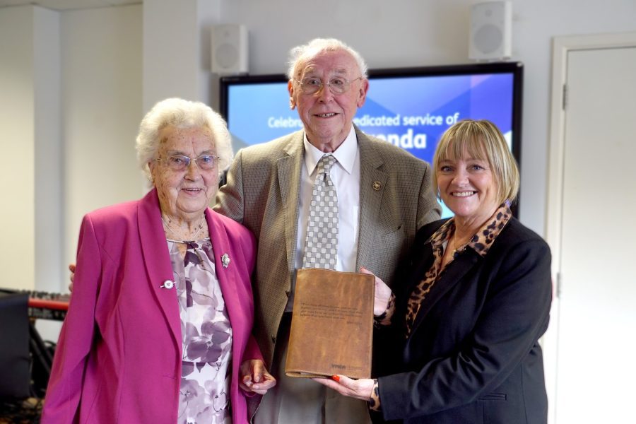 Eric and Brenda Moore smiling to camera, whilst being presented with a YMCA bible by CEO of YMCA England & Wales Denise Hatton recognising their years of service.