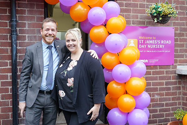 Man and woman staff team from YMCA stood smiling to camera below an orange and purple balloon arch at the grand opening of their new nursery in Dudley