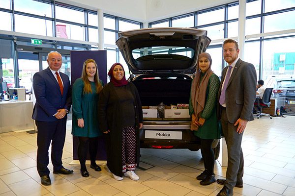 A group of people standing around a Vauxhall Mokka in a car showroom. The boot of the car is full of useful items to donate to families in need of a helping hand in Wolverhampton.