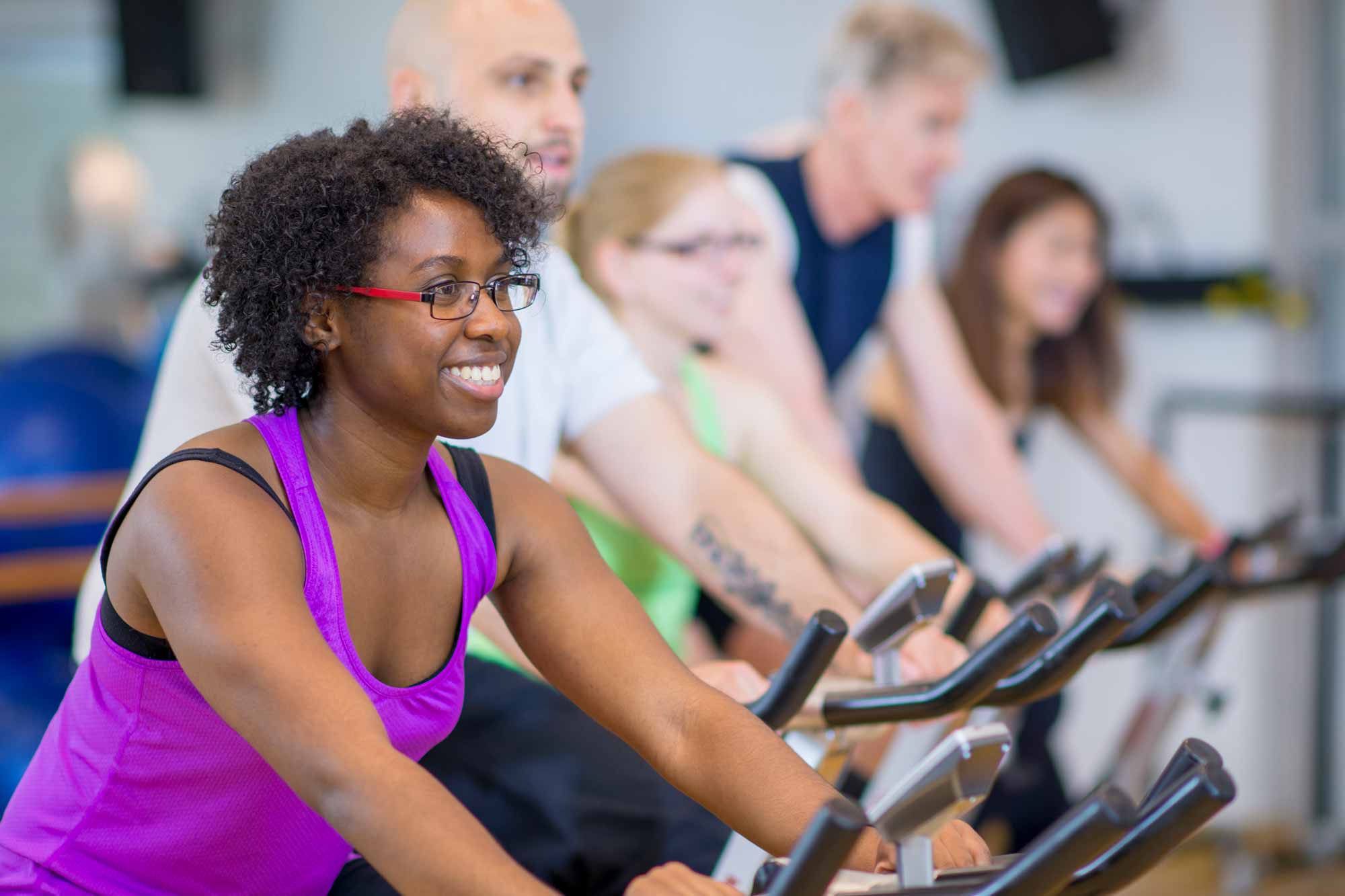 Group of people enjoying a spin class, lady in focus at the front smiling.
