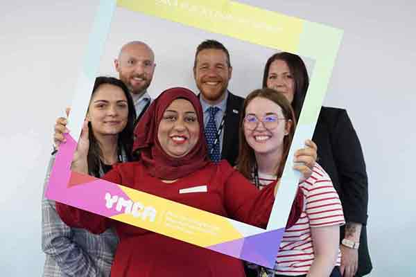 A group of diverse ymca staff members holding a selfie frame and smiling about their recent Gold Investors in People award.